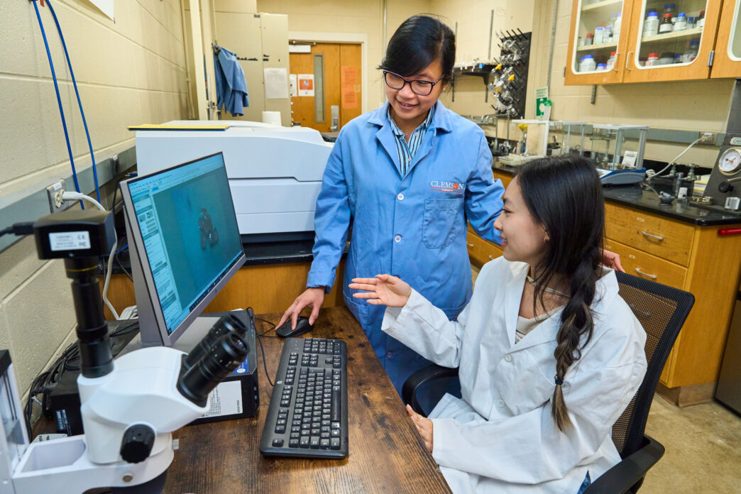 A woman wearing a blue lab coat (Thao Tran Dominy) listens to a student wearing a white lab coat while sitting in front of a computer.