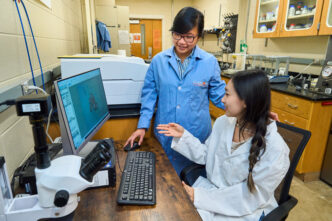 A woman wearing a blue lab coat (Thao Tran Dominy) listens to a student wearing a white lab coat while sitting in front of a computer.