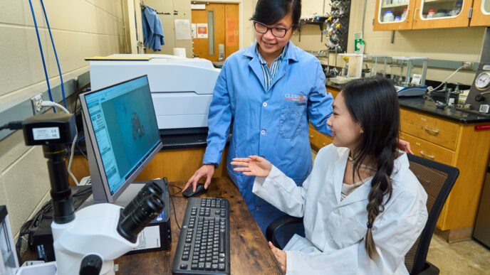 A woman wearing a blue lab coat (Thao Tran Dominy) listens to a student wearing a white lab coat while sitting in front of a computer.
