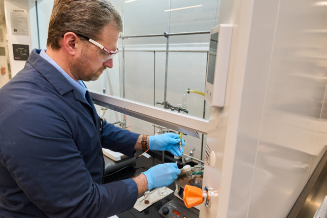 A man in a blue lab coat and wearing goggles works with something in a vent hood in a Clemson science lab.