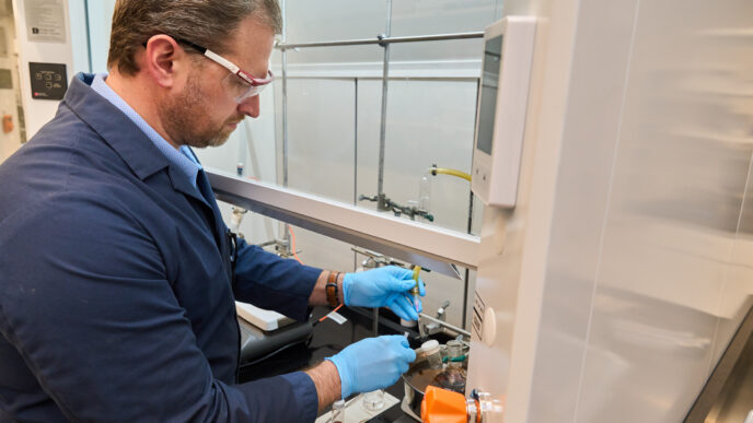 A man in a blue lab coat and wearing goggles works with something in a vent hood in a Clemson science lab.