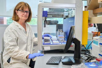 A woman with reddish brown hair wearing glasses sits in front of a big glass booth with scientific instruments inside of it.