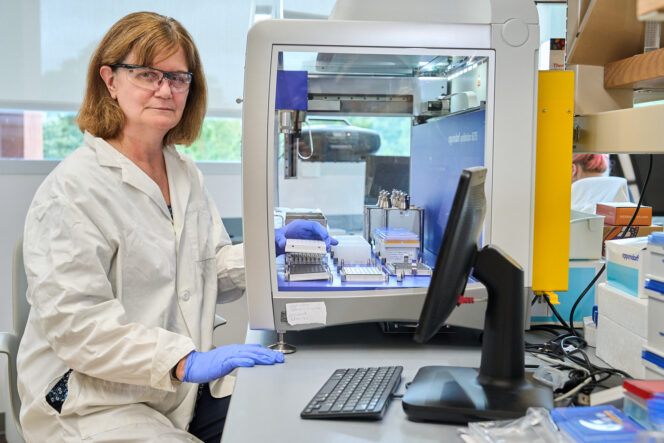 A woman with reddish brown hair wearing glasses sits in front of a big glass booth with scientific instruments inside of it.