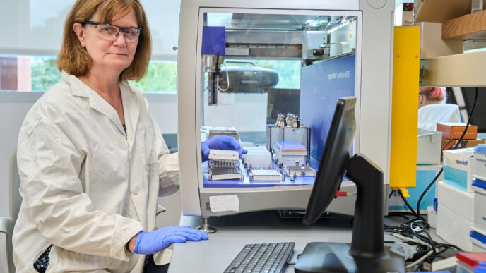A woman with reddish brown hair wearing glasses sits in front of a big glass booth with scientific instruments inside of it.