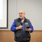 A man in a purple shirt and gray vest speaks with a microphone