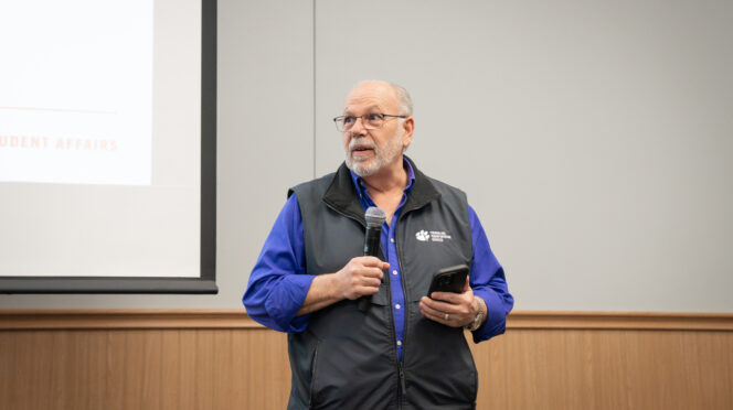 A man in a purple shirt and gray vest speaks with a microphone