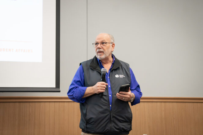 A man in a purple shirt and gray vest speaks with a microphone