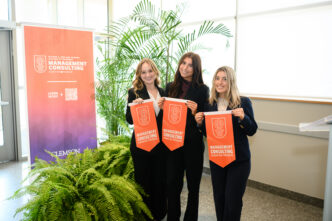 Three female students in black suits hold orange felt pennants in front of pop-up banner