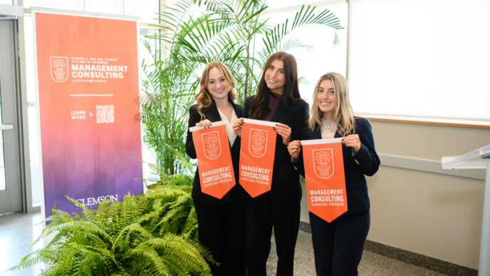 Three female students in black suits hold orange felt pennants in front of pop-up banner