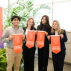 Three female students in black suits and one male student hold orange felt pennants in front of pop-up banner