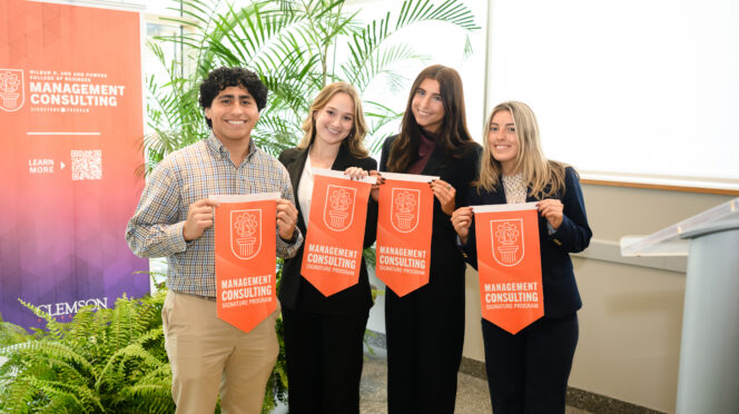 Three female students in black suits and one male student hold orange felt pennants in front of pop-up banner
