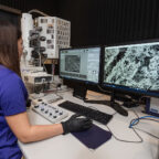 A woman wearing black safety gloves looks at a computer display showing images generated using electron microscopy.