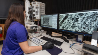 A woman wearing black safety gloves looks at a computer display showing images generated using electron microscopy.