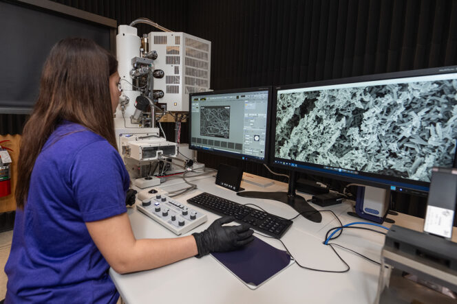 A woman wearing black safety gloves looks at a computer display showing images generated using electron microscopy.