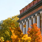 The front of a columned brick building that reads "Clemson University" at the top. Trees with fall leaves are in the foreground. leaves