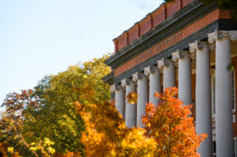 The front of a columned brick building that reads "Clemson University" at the top. Trees with fall leaves are in the foreground. leaves