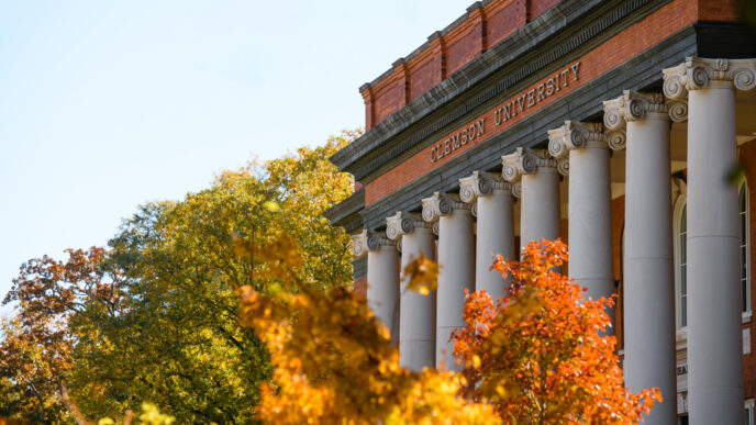 The front of a columned brick building that reads "Clemson University" at the top. Trees with fall leaves are in the foreground. leaves