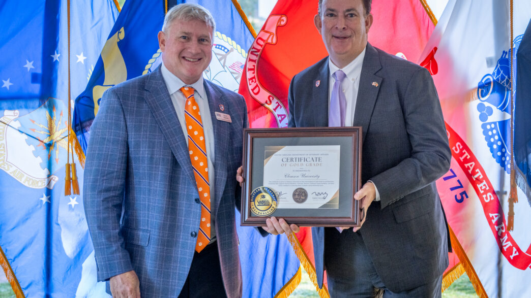 Two men in suits holding a plaque in front of military flags