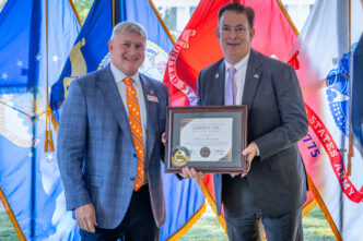 Two men in suits holding a plaque in front of military flags