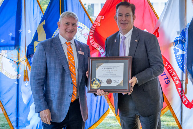 Two men in suits holding a plaque in front of military flags