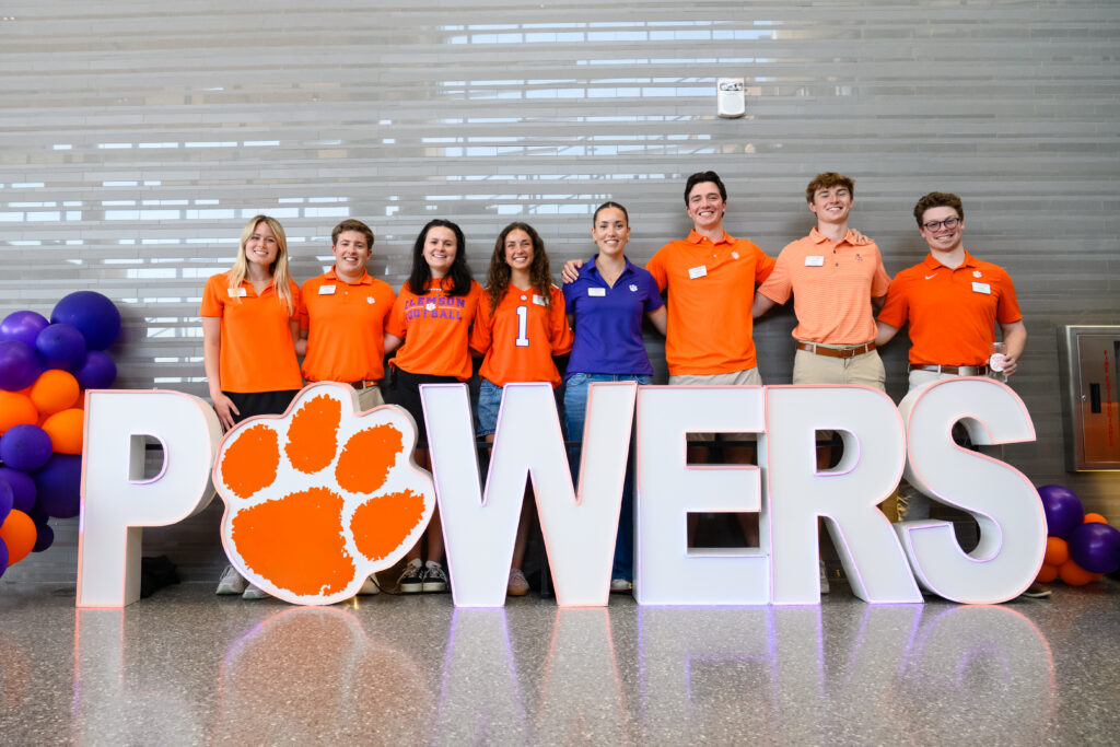 Eight business student ambassadors wearing orange Clemson spirit attire pose behind a large “POWERS” sign at a Wilbur O. and Ann Powers College of Business admitted student preview event.