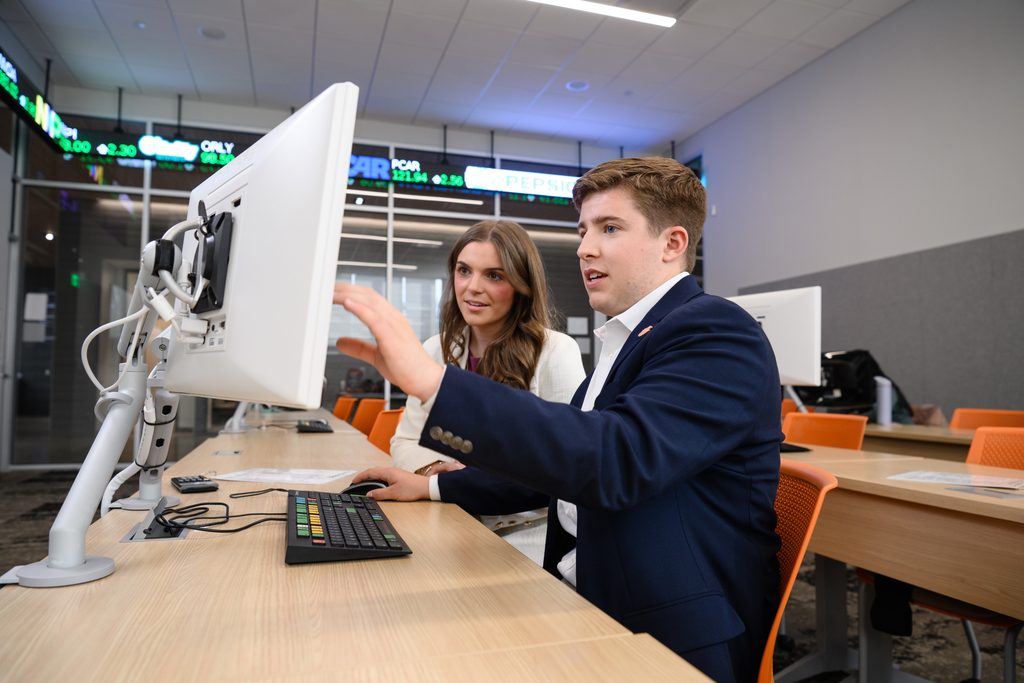 Male and female Clemson business students in professional attire look at a computer monitor inside the Melvin and Dollie Younts Trading Room.