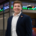 A male student wearing a navy blazer, khaki pants and a white button-down shirt stands inside a campus building with a stock trading ticker displayed in the background.