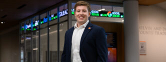 A male student wearing a navy blazer, khaki pants and a white button-down shirt stands inside a campus building with a stock trading ticker displayed in the background.