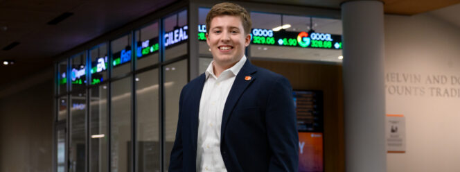 A male student wearing a navy blazer, khaki pants and a white button-down shirt stands inside a campus building with a stock trading ticker displayed in the background.