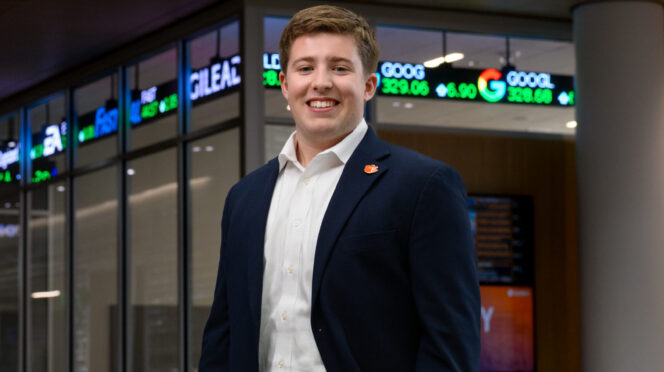 A male student wearing a navy blazer, khaki pants and a white button-down shirt stands inside a campus building with a stock trading ticker displayed in the background.