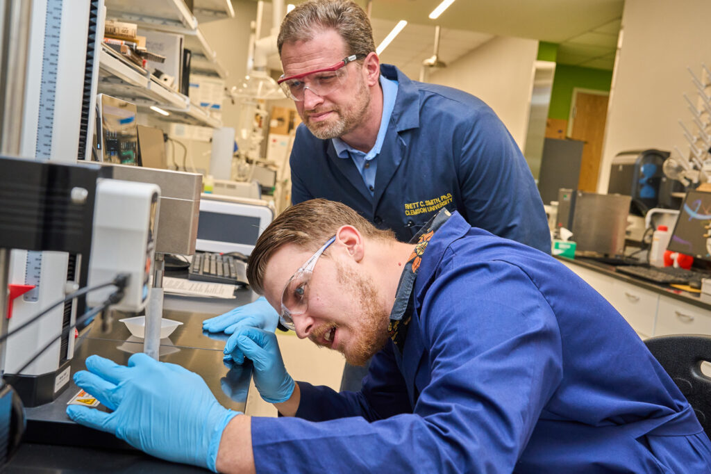 A man wearing a blue lab coat looks at a device in a chemistry lab while another man wearing a blue lab coat watches.