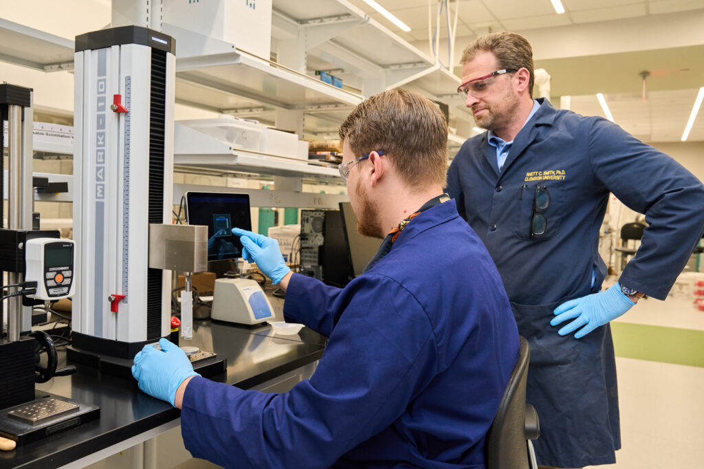 A man wearing a blue lab coat programs something into a machine while another man wearing a blue lab coat watches.